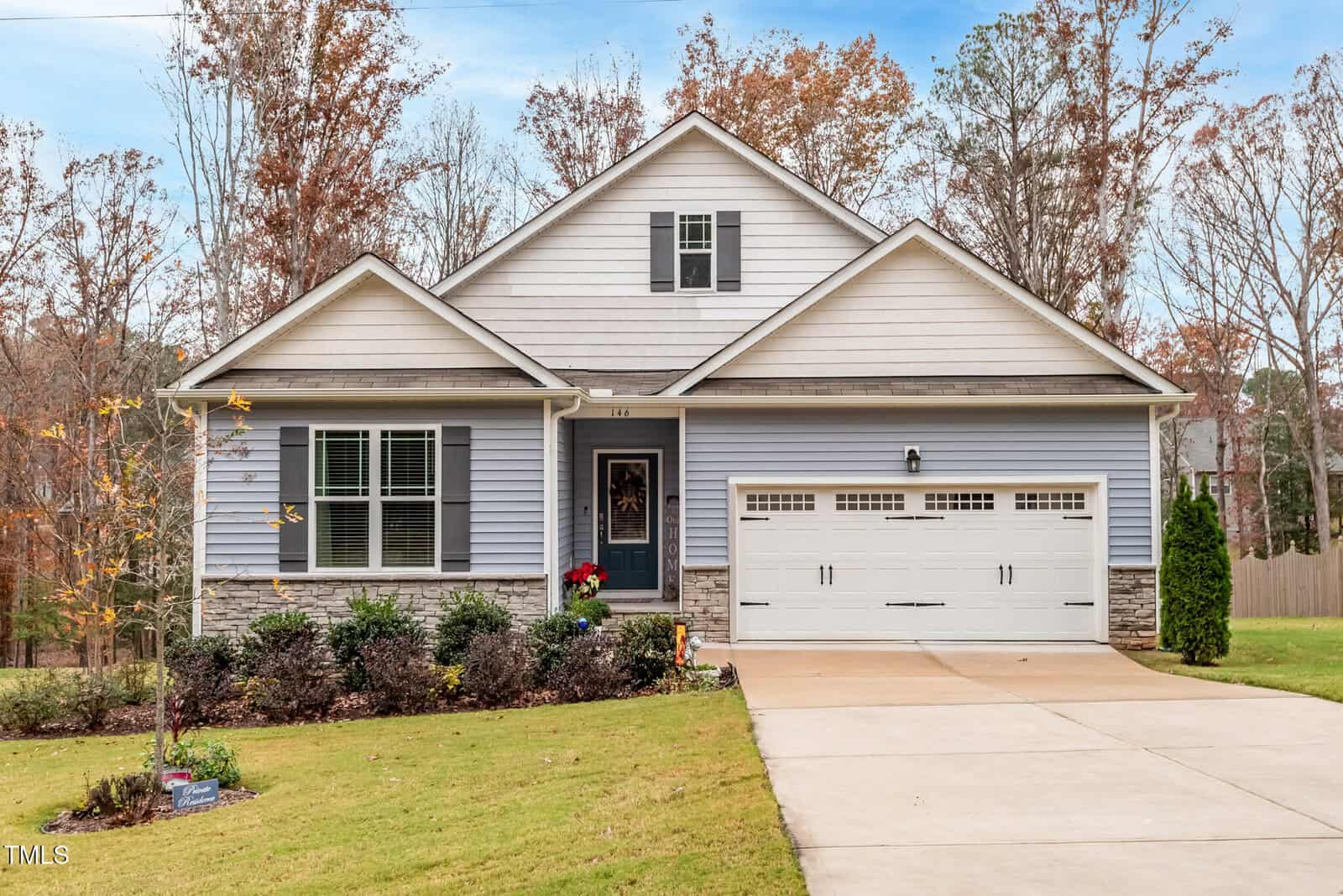 A single-story light gray house with white trim, a gable roof, and an attached two-car garage. The front yard is landscaped with shrubs and a lawn. Trees with autumn foliage stand in the background.
