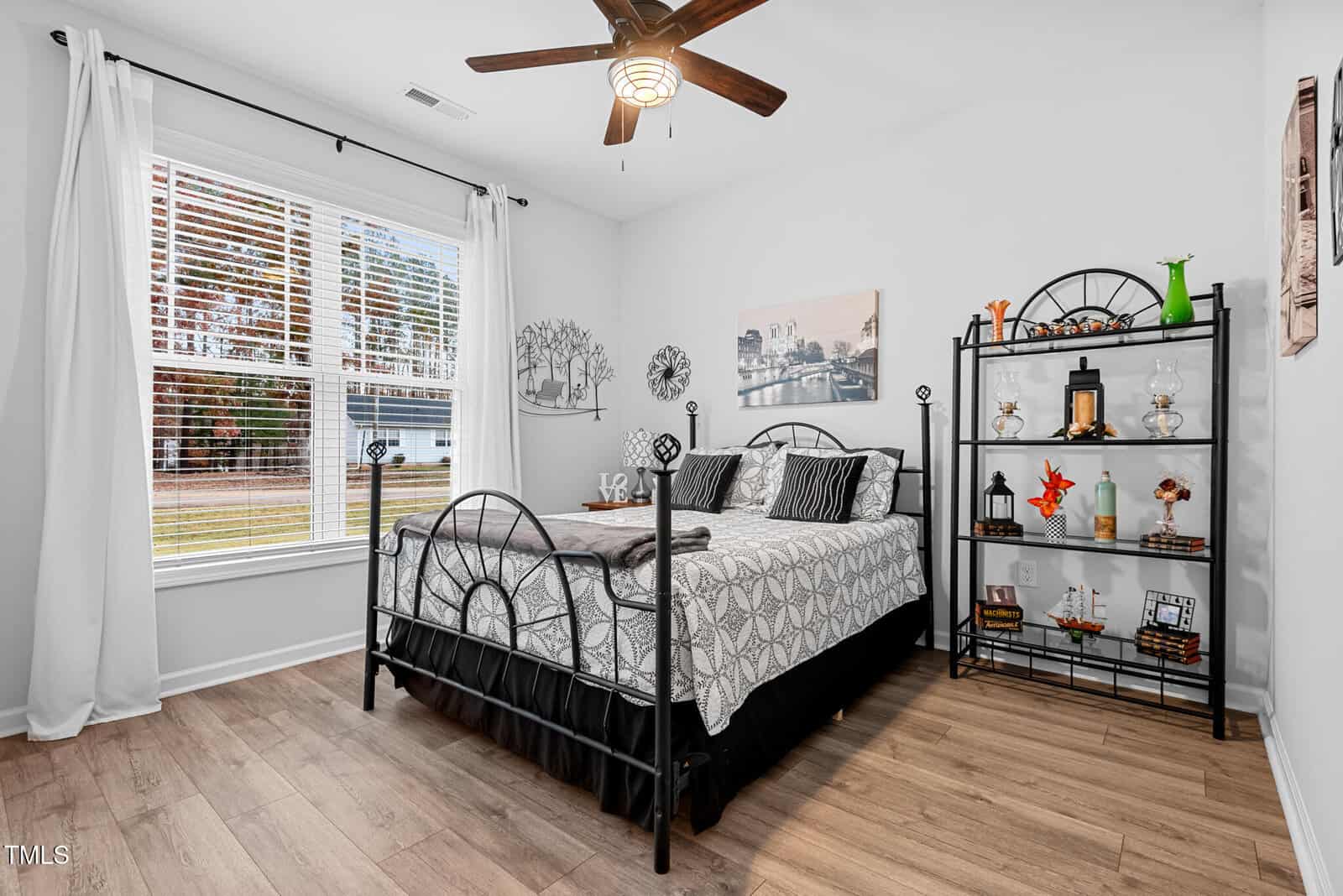 A bedroom with a metal-framed bed, geometric-patterned bedding, a black metal shelf displaying decorative items, wood flooring, a ceiling fan, and a large window with white curtains.