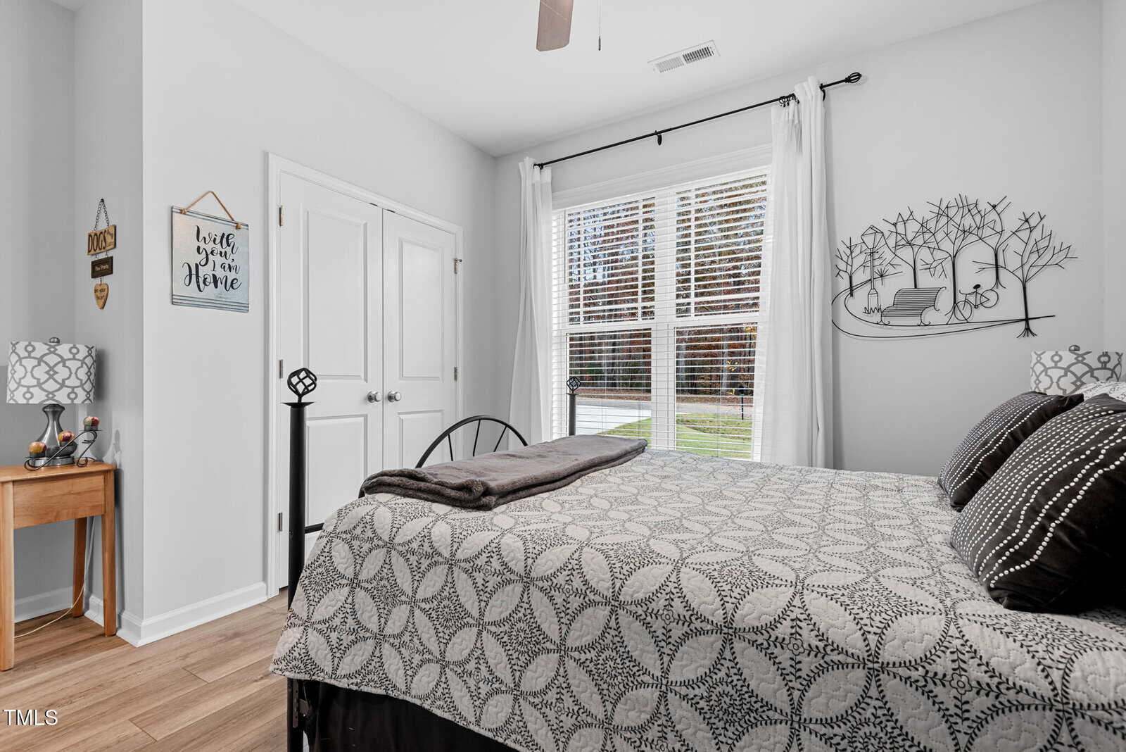 A bedroom with white walls, a bed with a gray patterned comforter, two matching bedside tables with lamps, a small wooden desk, white double doors, and a large window with white curtains letting in natural light.