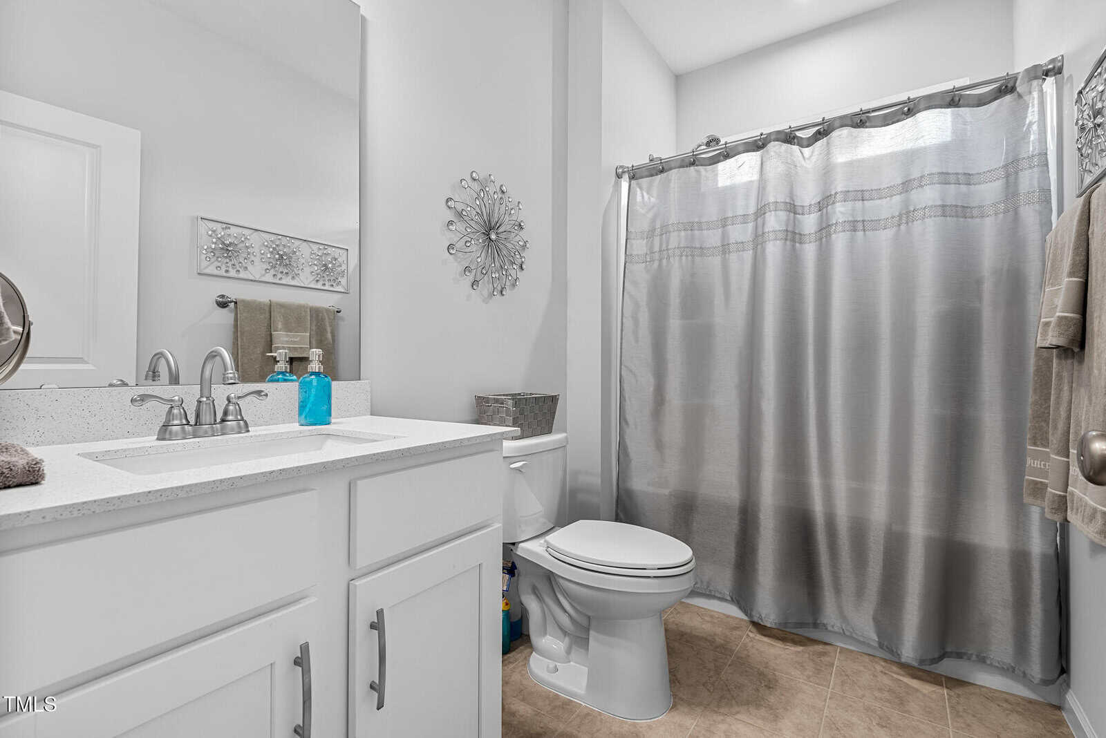 Modern bathroom with white cabinets, a sink, soap dispenser, toilet, gray shower curtain, towels on the wall, and decorative accents on light-colored walls and tile floor.