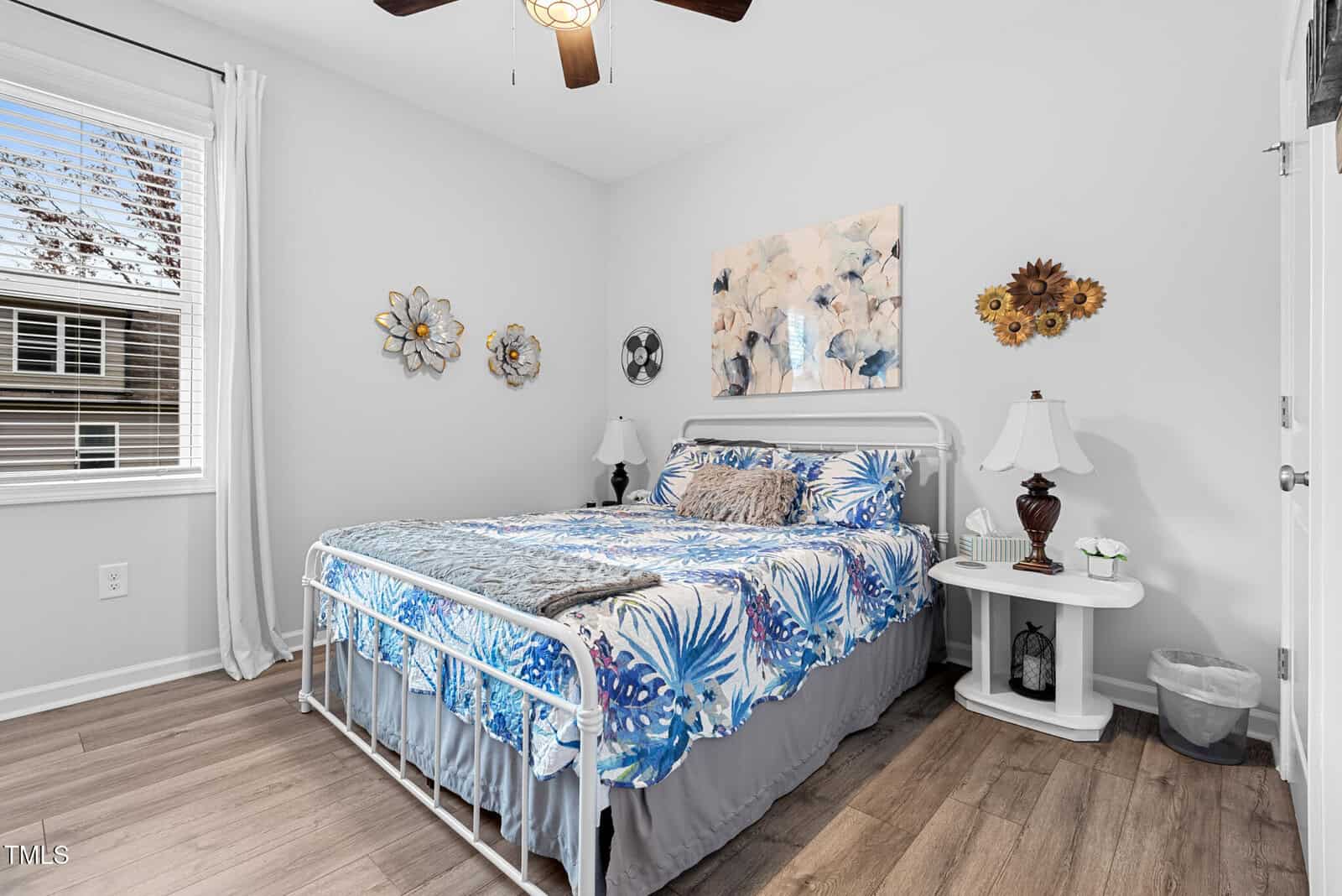 A cozy bedroom with a white metal bed, blue floral bedding, white nightstands with lamps, wall art, and a window letting in natural light. The room has light wood flooring and neutral walls.