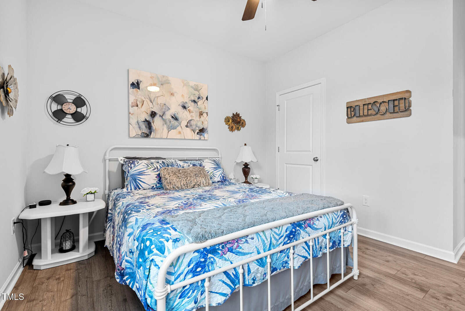 A bedroom with a white metal bed frame, blue floral bedding, two bedside tables with lamps, a ceiling fan, and wall decor including a “BLESSED” sign and floral art. The room has white walls and wood flooring.