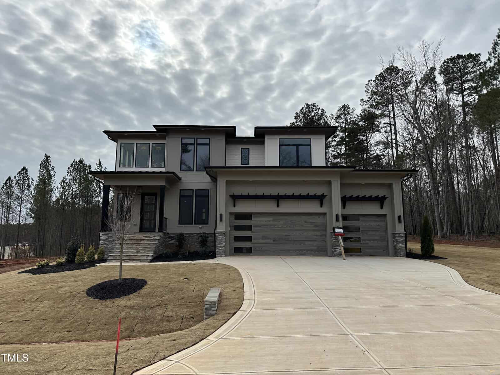 A modern two-story house with light and dark exterior finishes, large windows, a three-car garage, and a wide driveway, surrounded by bare trees and landscaped yard under a cloudy sky.
