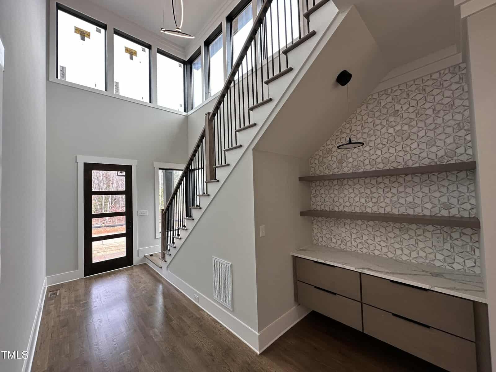 A modern entryway with large windows, wood flooring, a wooden door with glass panels, and a staircase with metal railings. Built-in shelves and drawers are under the stairs, with a geometric tile backsplash.