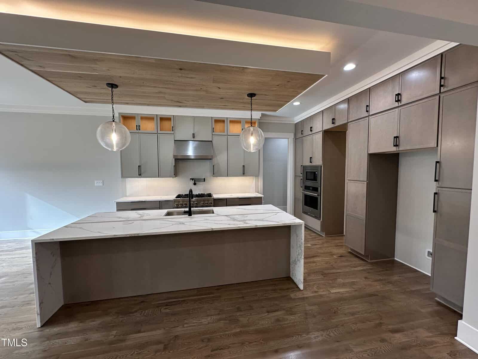 Modern kitchen with a marble island, wood floors, beige cabinetry, stainless steel appliances, two globe pendant lights, and a wood-paneled ceiling accent above the island.