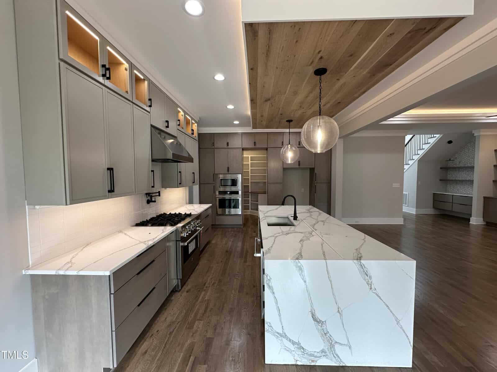 Modern kitchen with marble countertops, large island with sink, wood floors, gray cabinets, stainless steel appliances, decorative ceiling with wood panel, and two globe pendant lights. Open shelving and stairs visible in background.