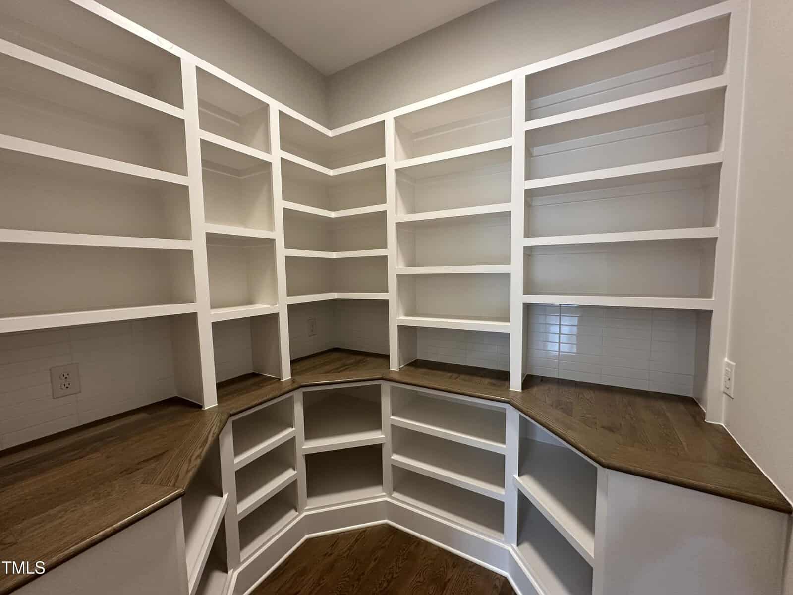Empty walk-in pantry with wooden countertops and white built-in shelves covering two walls, forming an L-shape. The floor is wood, and the walls are light-colored with a tile backsplash.