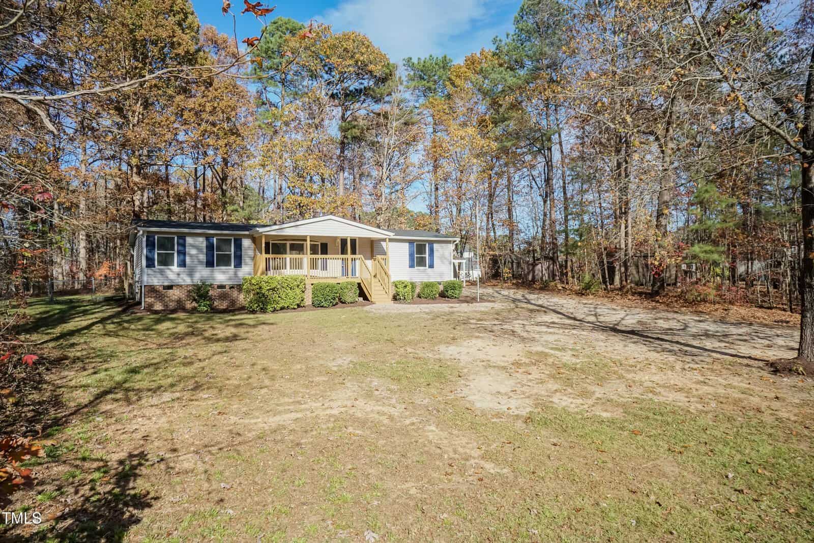A single-story house with a small front porch and steps, white siding, and green shutters, sits on a spacious lot with a dirt driveway, surrounded by trees with autumn foliage.
