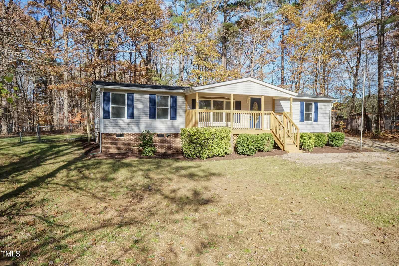 A single-story house with light-colored siding, blue shutters, and a small front porch with wooden railings and steps. The front yard has shrubs, grass, and is surrounded by tall trees with autumn foliage.