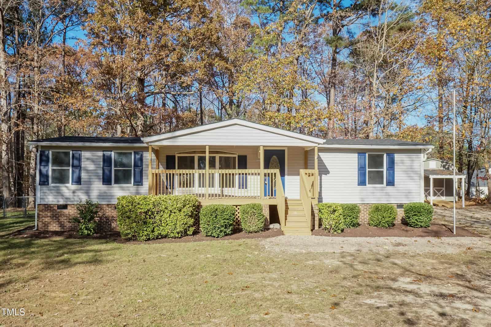 Single-story house with beige siding, brick foundation, and a covered front porch with steps. Bushes line the front of the house, and there are trees with autumn leaves in the background.