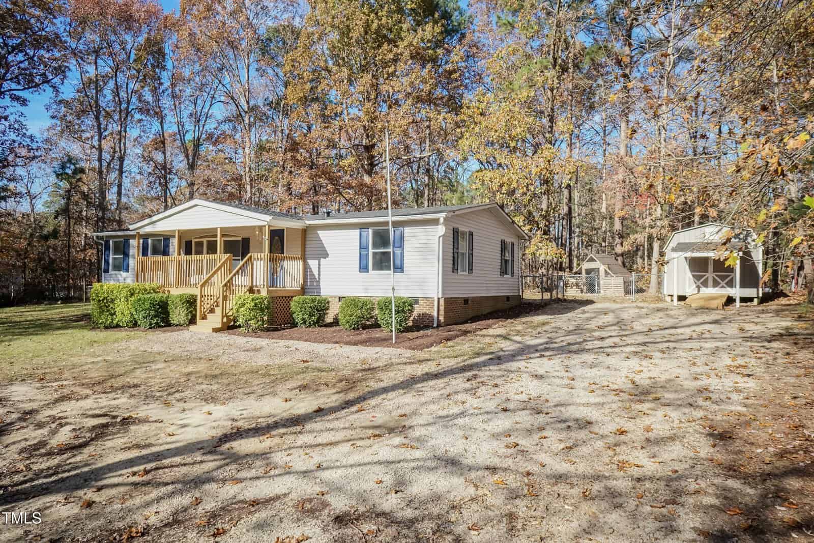 A single-story beige manufactured home with a small front porch sits on a dirt driveway, surrounded by autumn trees. A white shed is visible in the background to the right of the house.
