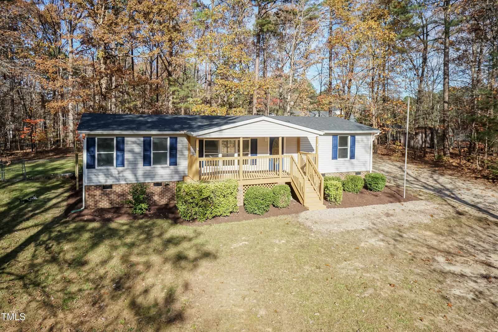 Single-story house with beige siding, brick foundation, and a small front porch with steps. Surrounded by bushes and trees with autumn foliage, and a grassy yard in front.