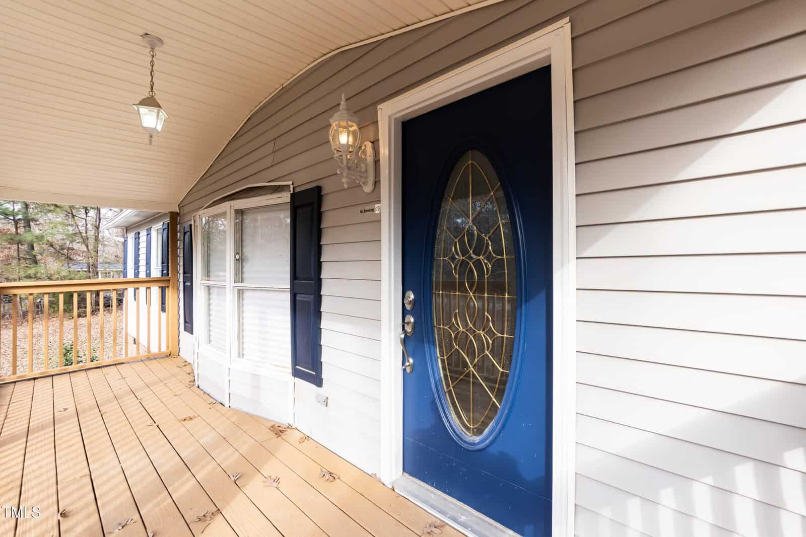 A close-up view of a house’s front porch with a blue oval glass door, beige siding, bay window, wooden floor, and hanging lights.
