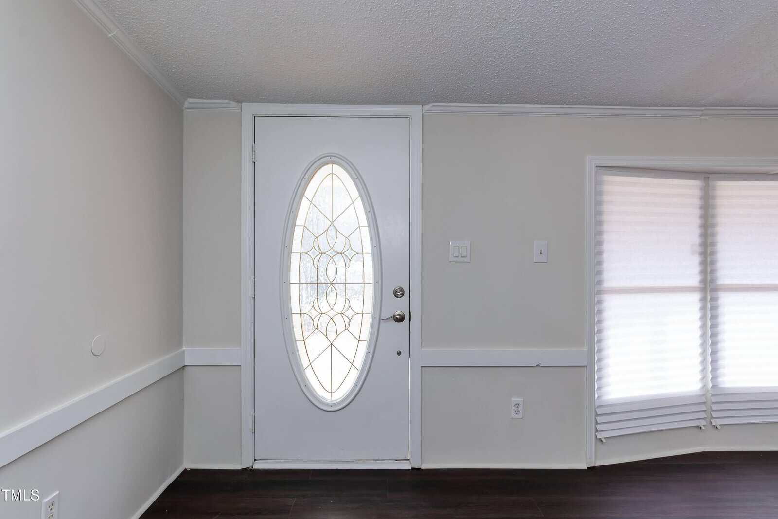 A white door with an oval glass window featuring a decorative pattern is centered in a light gray room with dark wood floors and a window with closed blinds to the right.