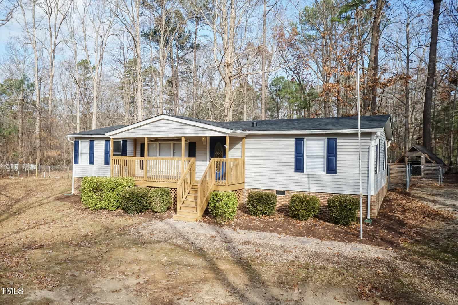 Single-story light-colored house with a covered front porch, blue shutters, and brick foundation. Wooden steps lead to the front door. Surrounded by trees and bushes, with a dirt yard in the foreground.