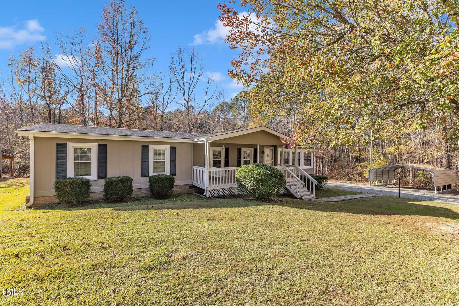A single-story beige house with a front porch is surrounded by a well-kept lawn and trees with fall foliage. A small driveway leads to a detached carport on the right. The sky is clear and blue.