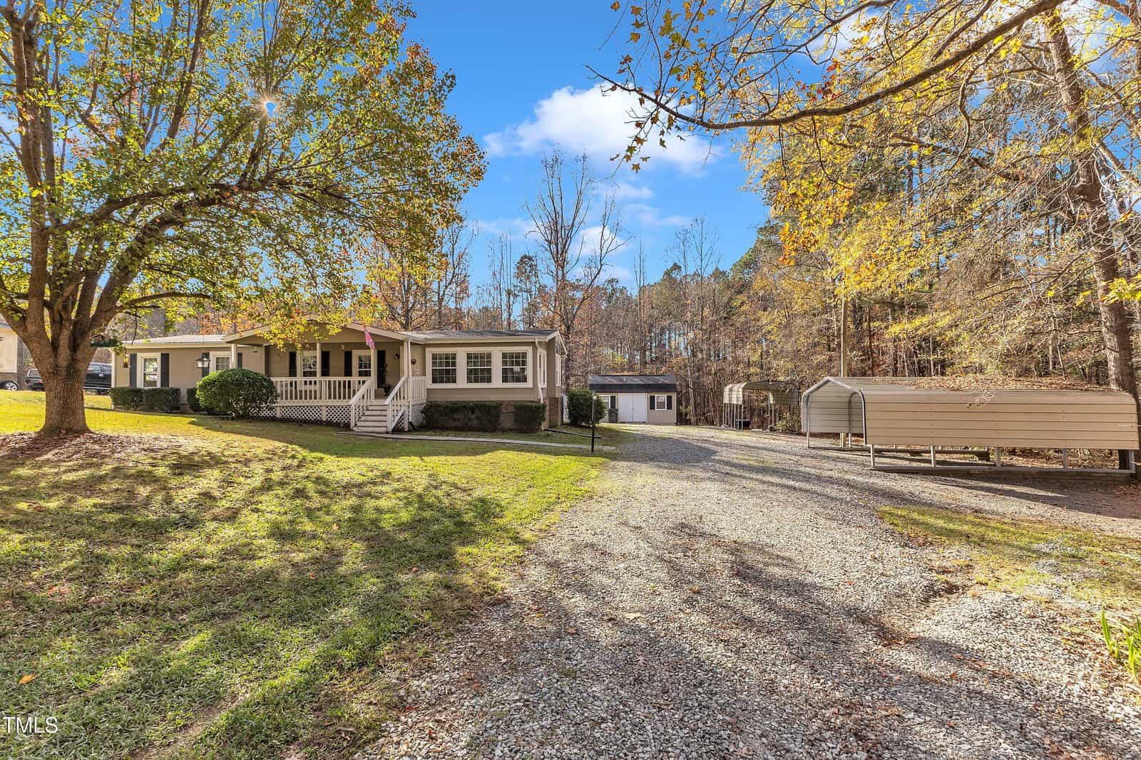 A single-story house with a porch is surrounded by trees with autumn foliage. A gravel driveway leads to the house, and there is a small outbuilding or shed to the side. The sky is clear with a few clouds.