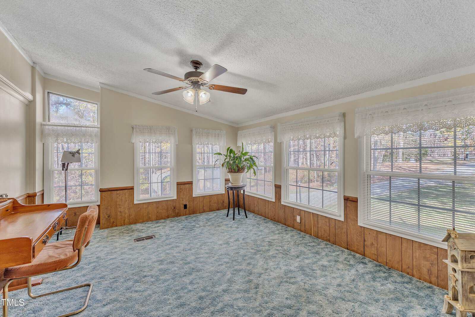 A bright sunroom with multiple windows, featuring a ceiling fan and blue carpet. The room includes a wooden desk with chairs on the left and a large potted plant in the center, with wood-paneling halfway up the walls.