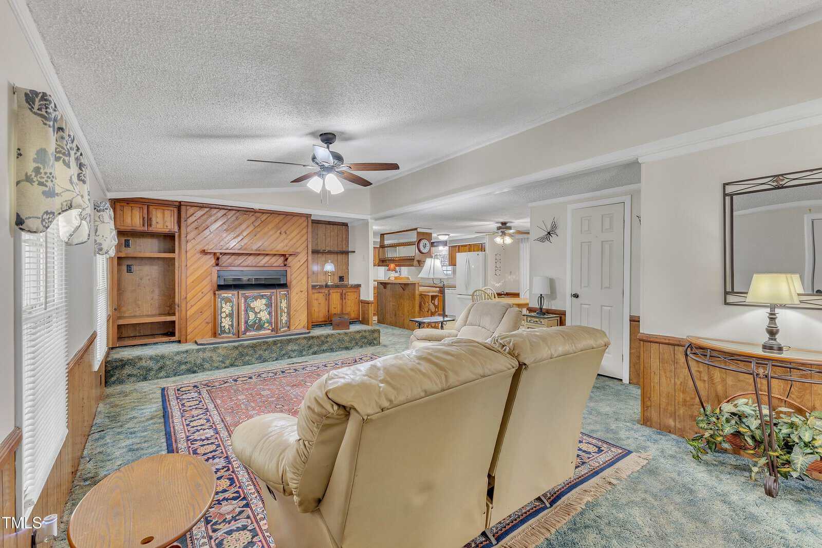 Spacious living room with beige carpet and brown wooden paneling. Features a large beige sofa, wooden coffee table, and a fireplace. Ceiling fans with lights are installed, and a small dining area is visible in the background. A large area rug is centered.