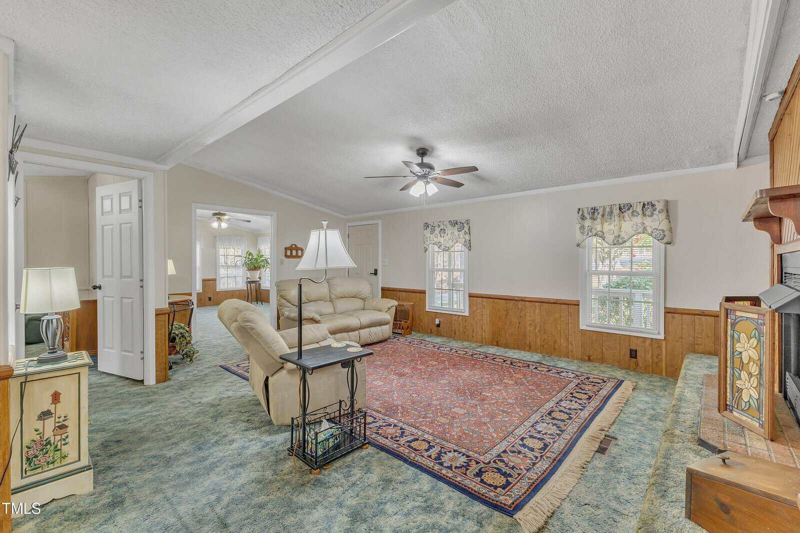 A living room with a blue-green carpet and a large red patterned rug. It features a beige sofa, a floor lamp, and ceiling fans. There is a decorative fireplace on the right, and two windows with floral curtains along one wall. An open doorway leads to another room.
