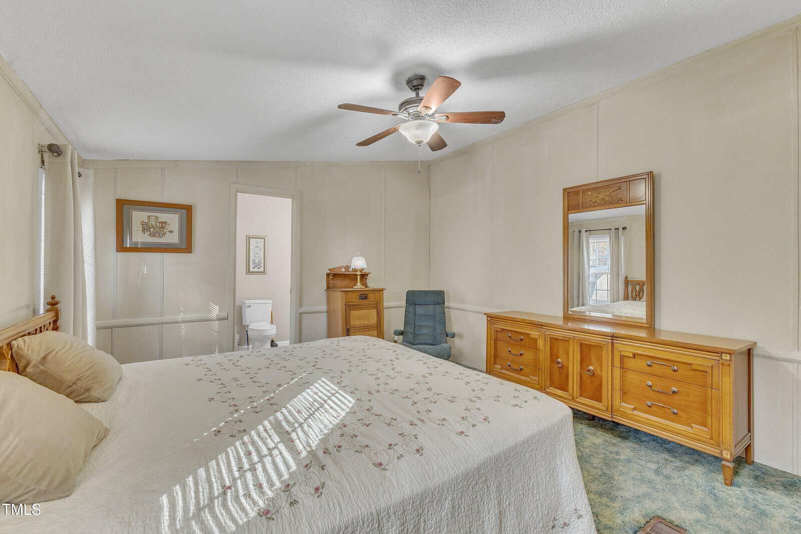 A bedroom featuring a large bed with a patterned quilt, a wooden dresser with a mirror, and a dark reclining chair. An overhead ceiling fan provides light, and an open door reveals a glimpse of a bathroom with a toilet.