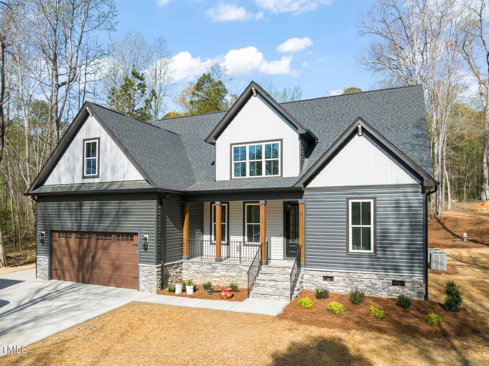 A modern two-story house with gray siding, white trim, and a dark shingled roof, featuring a double garage, front porch with wooden columns, and landscaped yard on a sunny day.
