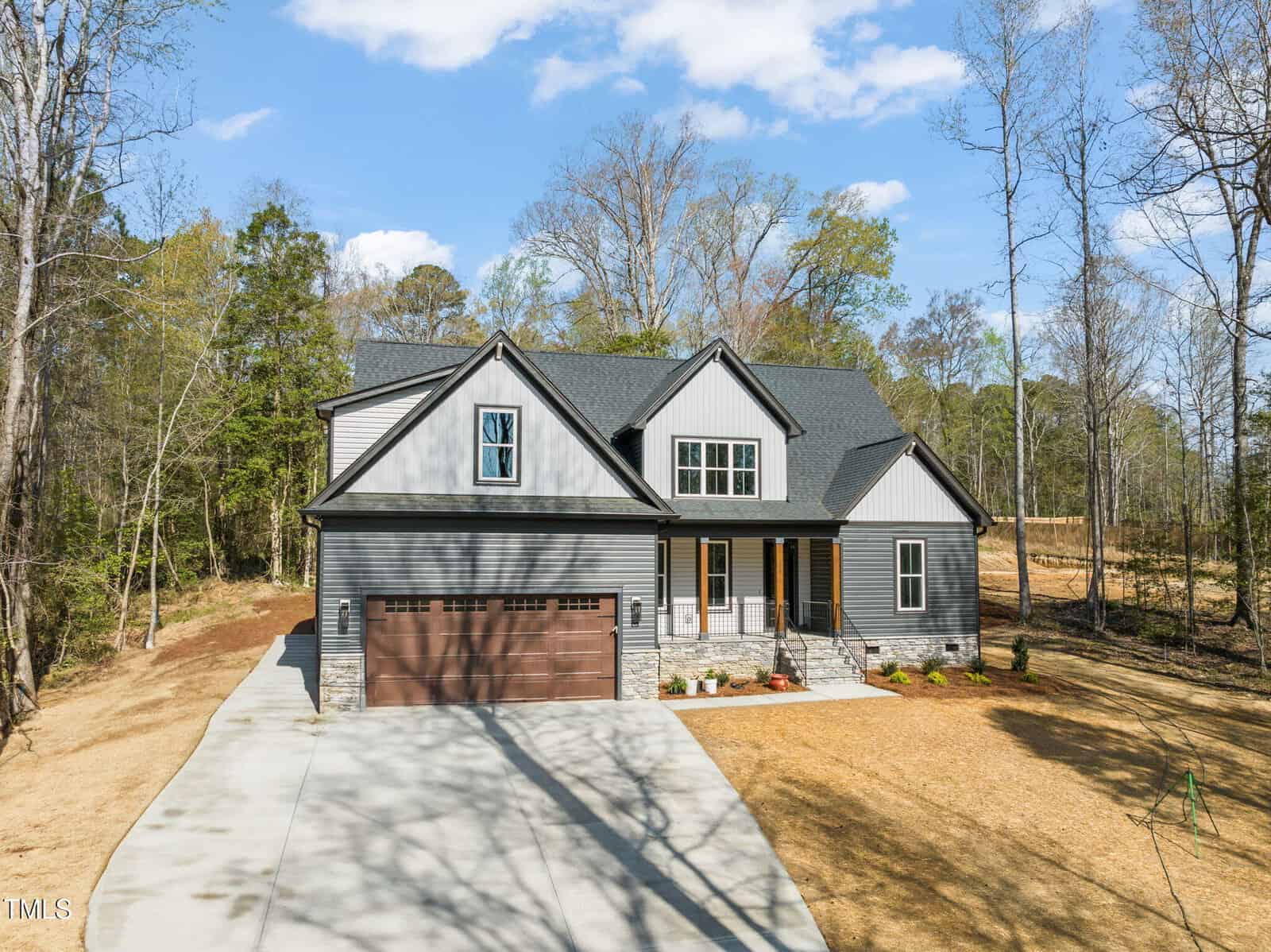 A modern gray house with white trim and a brown double garage sits at the end of a long concrete driveway, surrounded by trees and a sparse lawn under a partly cloudy sky.