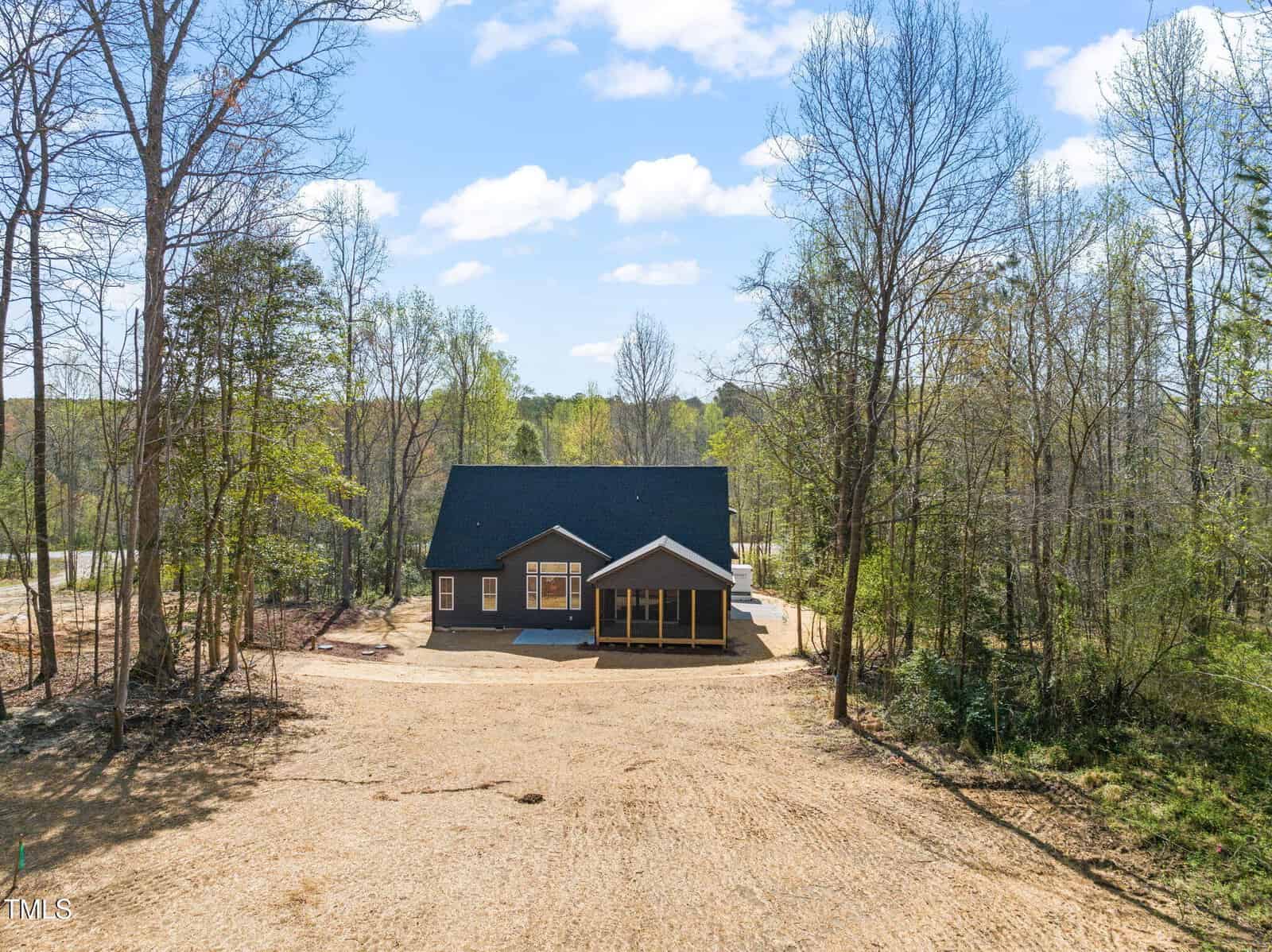 A single-story house with a dark exterior and a gable roof is situated in a clearing surrounded by trees, with a large, bare dirt yard in front under a partly cloudy sky.