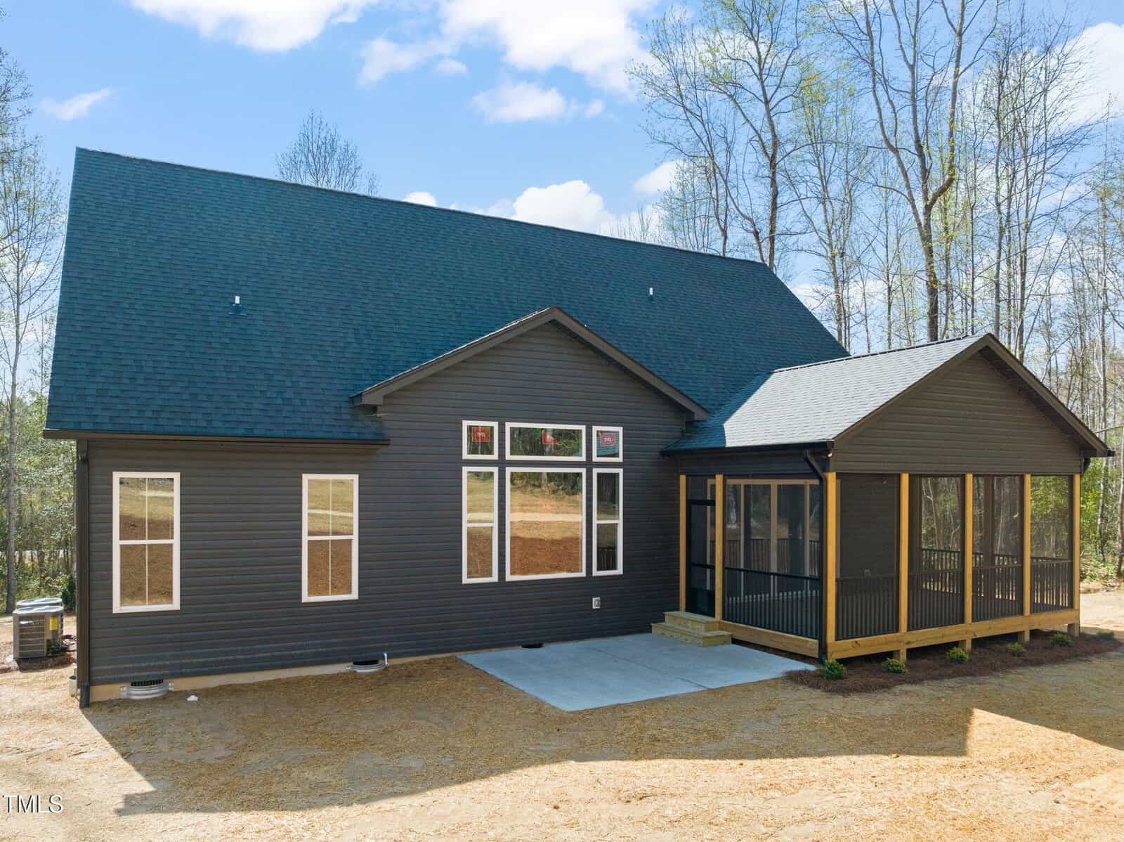 A single-story black house with large windows, a dark shingled roof, and a screened-in porch sits on a cleared lot surrounded by trees under a partly cloudy sky.