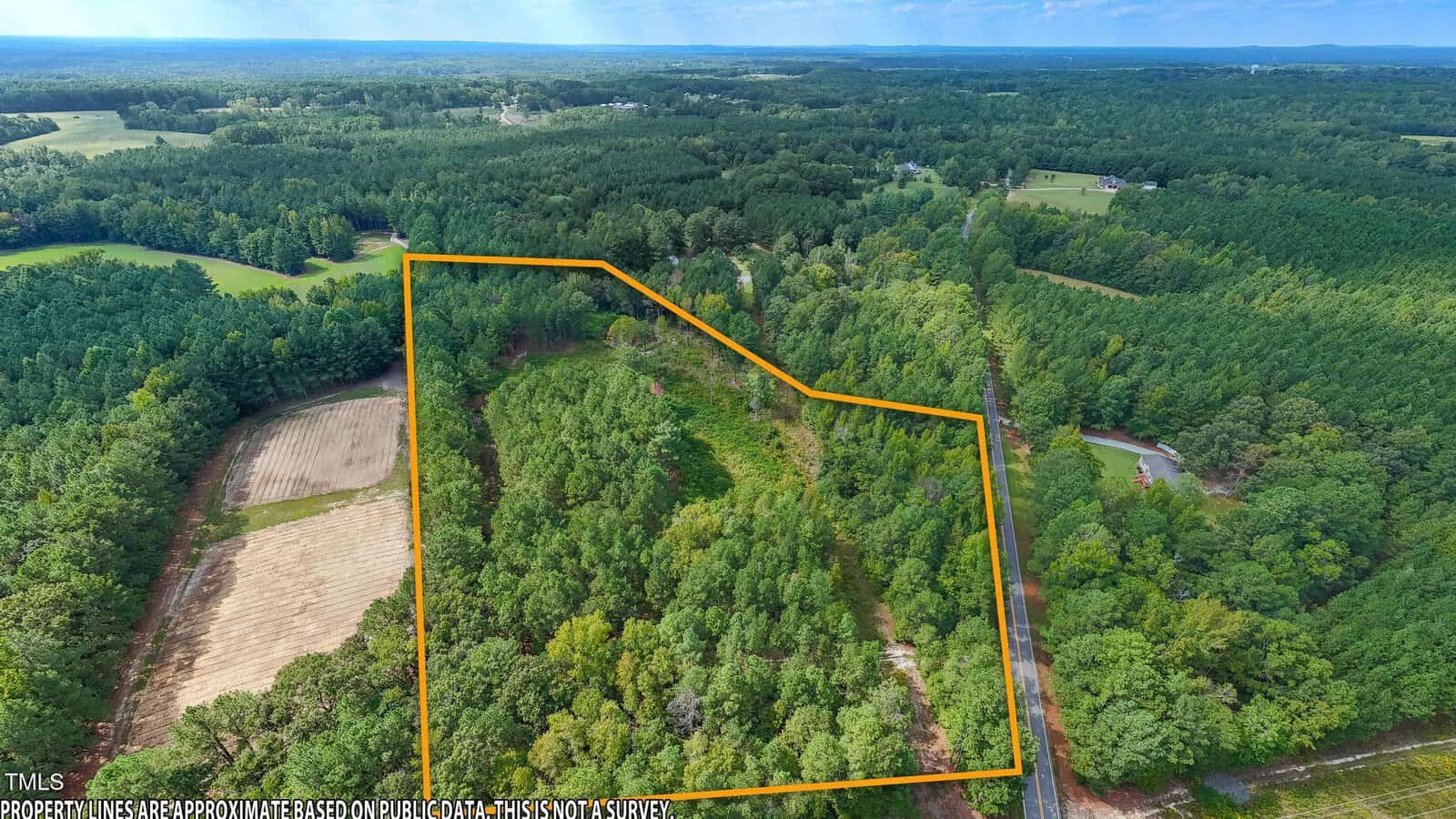 Aerial view of a densely forested area with the Lot 4 Dove Rd property boundary outlined in orange. Adjacent to this Creedmoor gem are open fields and a road that runs alongside part of the boundary. The horizon reveals a vast expanse of trees under a blue sky.