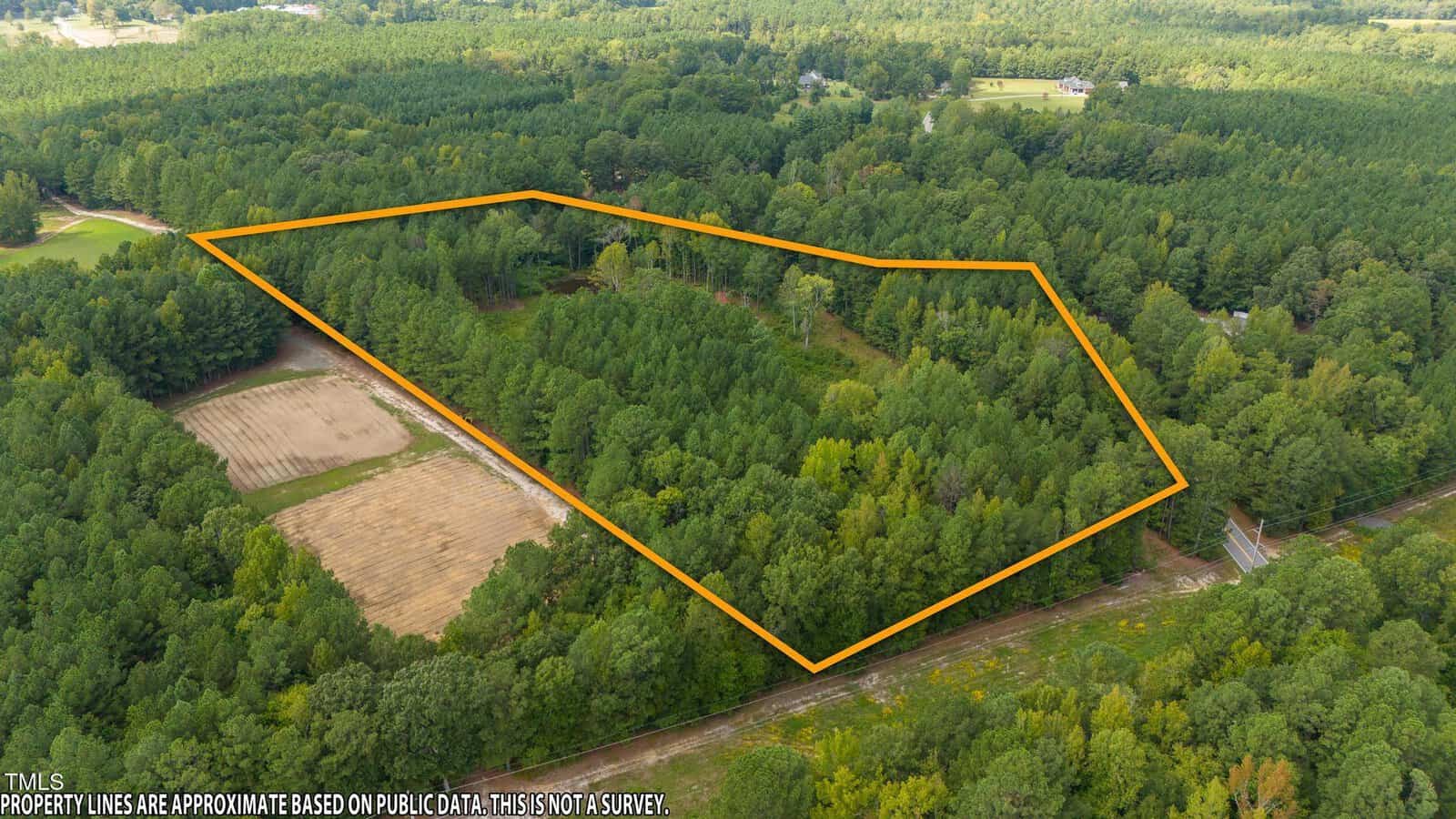 Aerial view of a densely wooded area outlined with an orange border, indicating a specific plot of land. Surrounding the plot are trees and patches of cleared land, with a few structures visible in the distance.