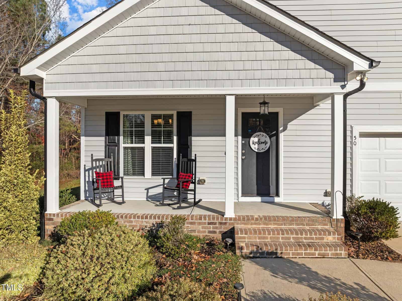 A front porch with gray siding, white trim, two black rocking chairs with red plaid cushions, a hanging lantern, and a round welcome sign on the black front door. Bushes and a walkway are in front of the porch.