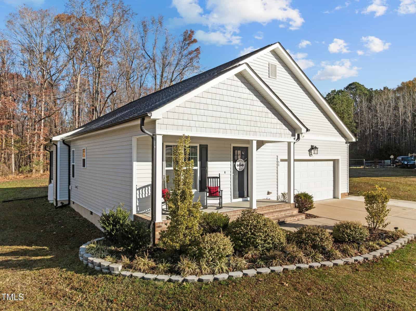 White single-story house with a two-car garage, dark roof, and small front porch. There are two red rocking chairs and neatly trimmed shrubs along a curved stone border. The yard is grassy with trees in the background.