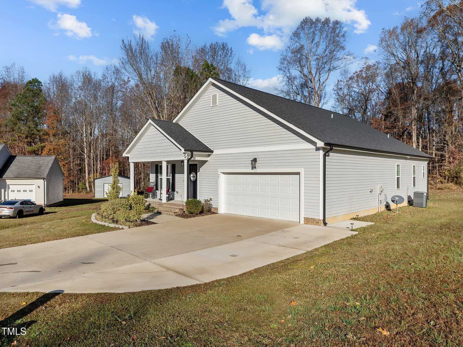 A light gray single-story house with a two-car garage, front porch, and sloped driveway sits on a grassy lawn, surrounded by trees under a blue sky with scattered clouds.