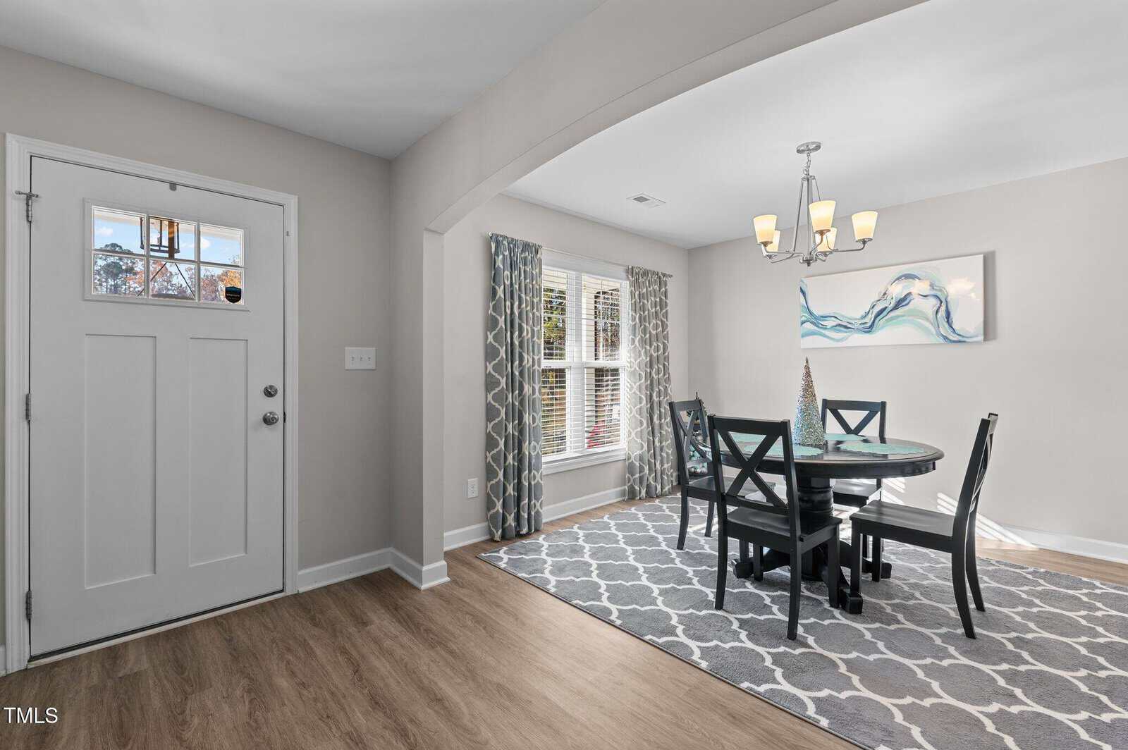 Bright dining room with a round table, four chairs, chandelier, large window with patterned curtains, abstract art, and a white front door, all on a gray patterned rug and wood flooring.