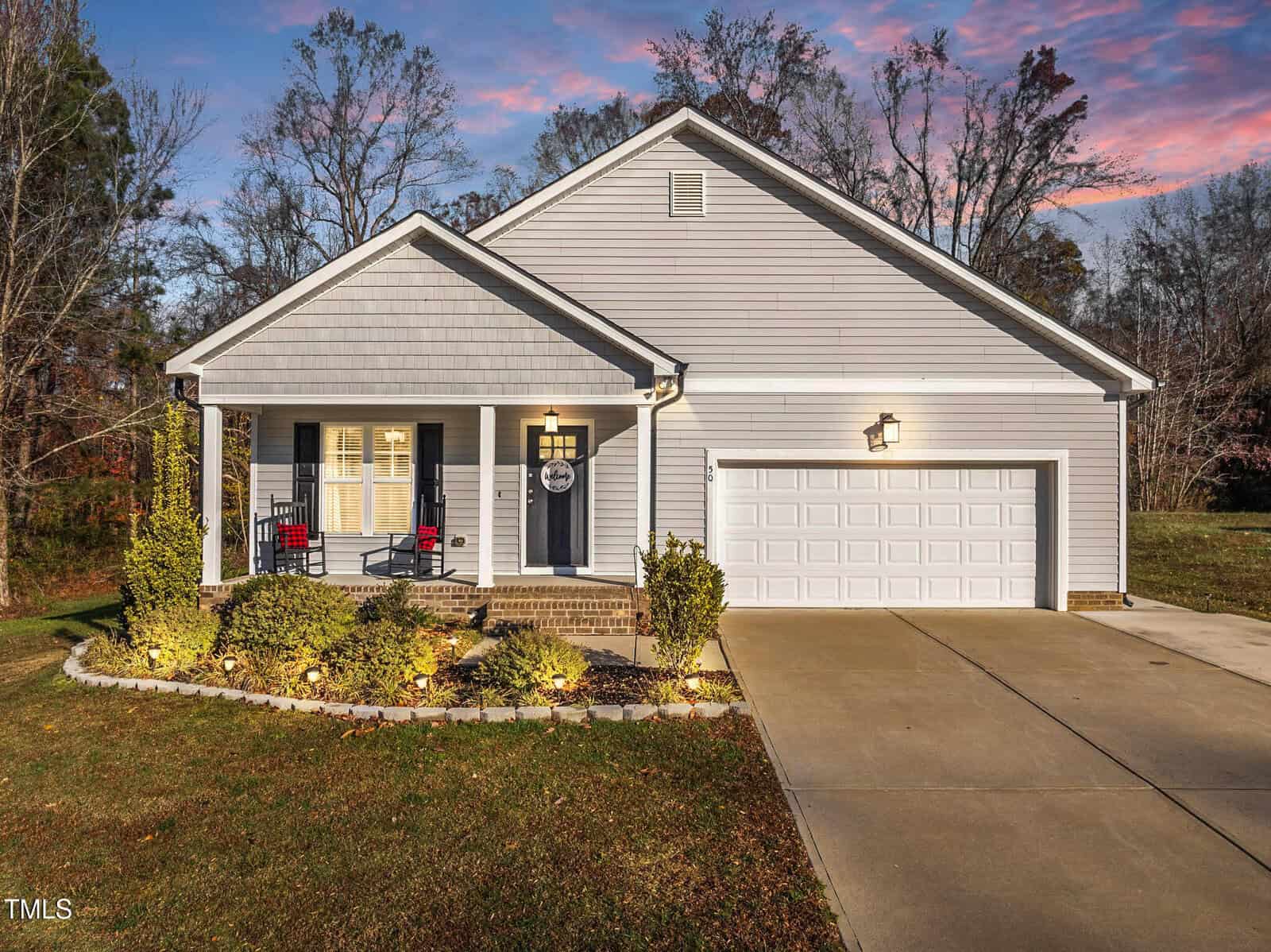 Single-story gray house with white trim, a covered front porch with two red chairs, a white garage door, and a concrete driveway. The yard is landscaped with shrubs and surrounded by trees at sunset.