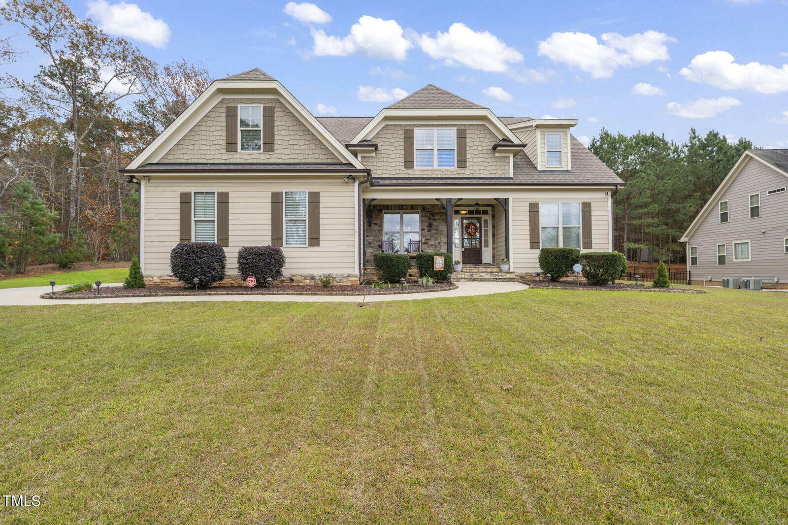 A two-story beige suburban house with stone accents, black shutters, and a covered front porch, surrounded by trimmed bushes and a large, neatly mowed yard under a blue sky with scattered clouds.