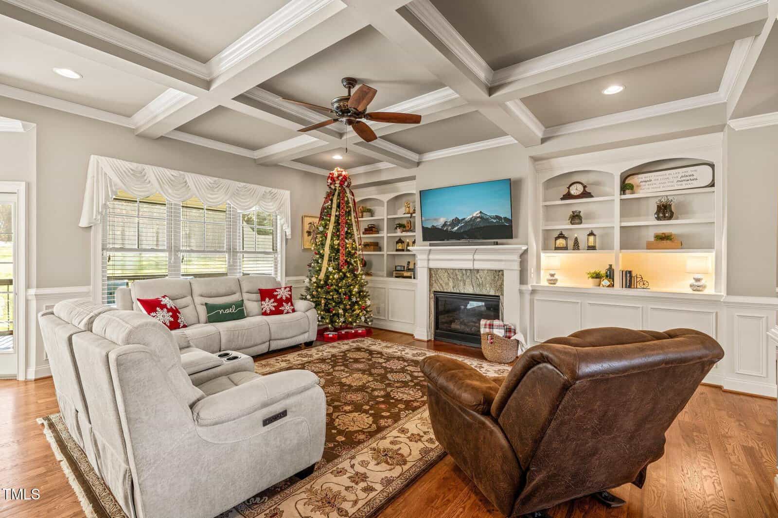 A cozy living room with a Christmas tree, beige sofas, a brown recliner, built-in shelves, a rug, a ceiling fan, and a TV above a fireplace. Natural light streams in through large windows with white curtains.