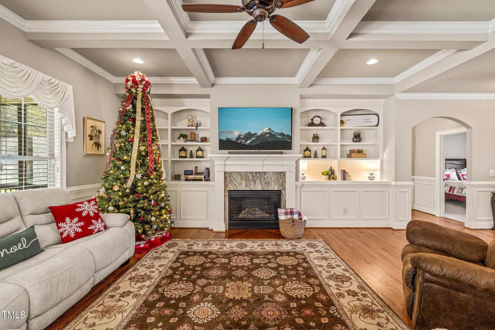 A living room features a decorated Christmas tree, a white fireplace with a TV above it, built-in shelves with decor, a beige sofa, a brown recliner, and a patterned rug on a wooden floor.