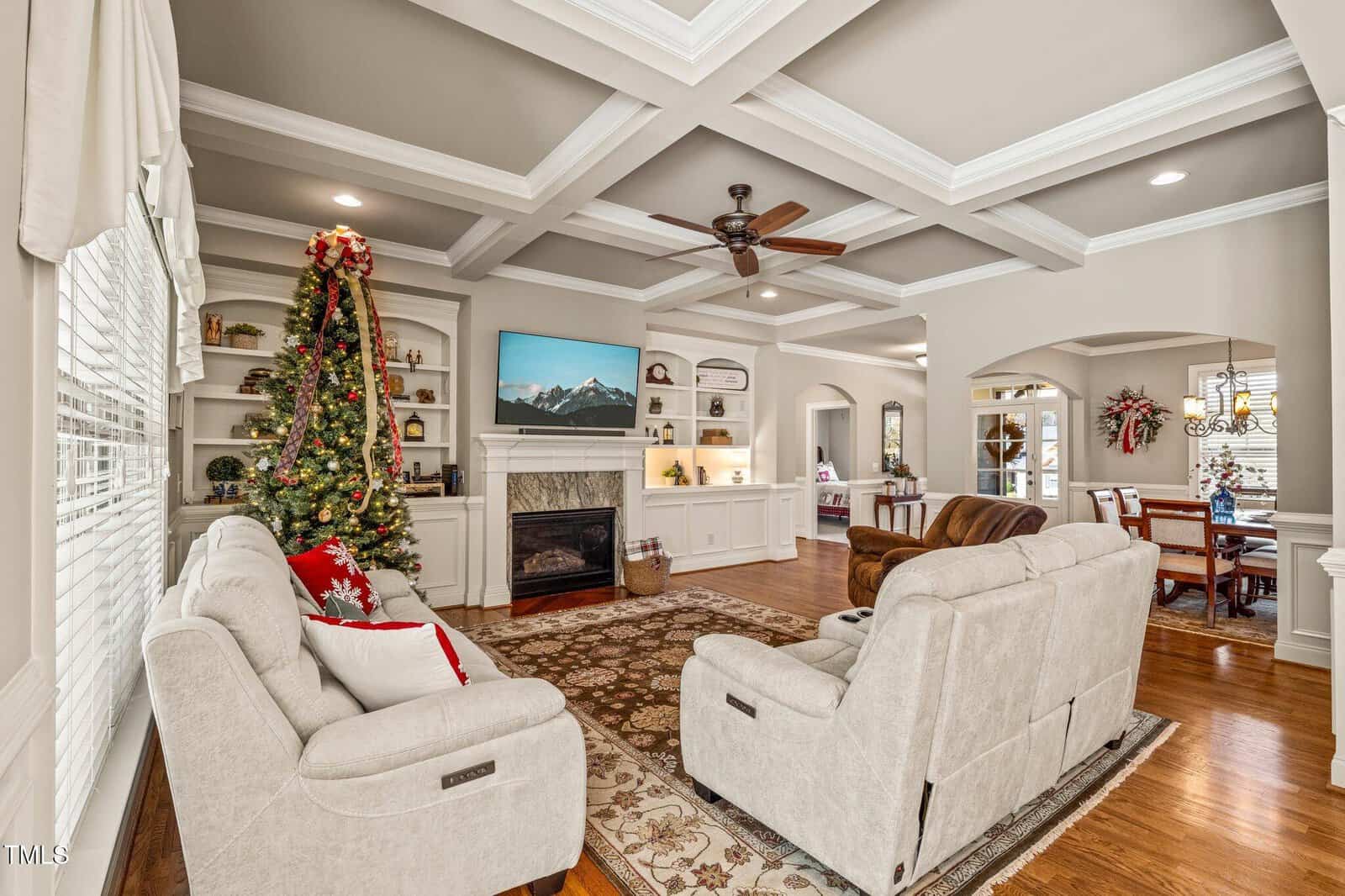 A cozy living room with beige sofas, a decorated Christmas tree, a fireplace, TV, built-in shelves, and wood flooring. The adjacent dining area is visible, featuring a festive wreath.