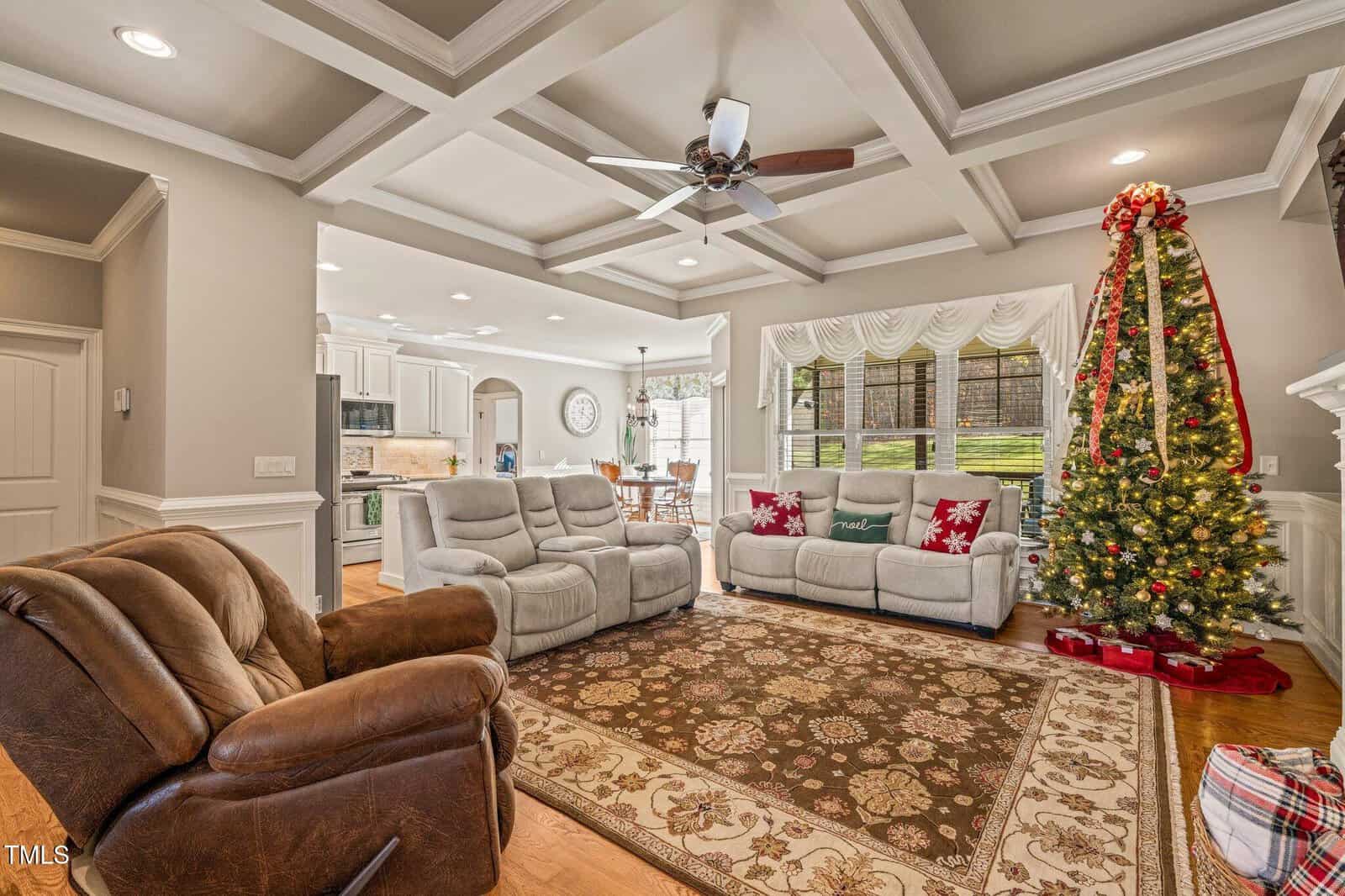 A living room with a brown recliner, two gray sofas, a patterned area rug, and a decorated Christmas tree with wrapped gifts. The room opens to a kitchen and dining area with white cabinets and wood flooring.