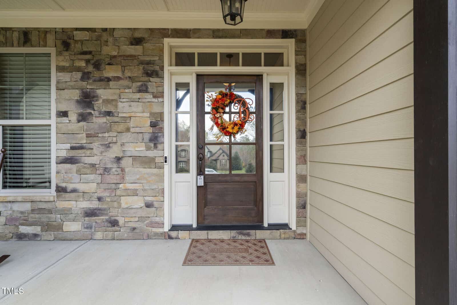 A front porch with a stone and siding exterior features a wooden door with glass panels, a fall-themed wreath, a doormat, and a light fixture above the entrance.