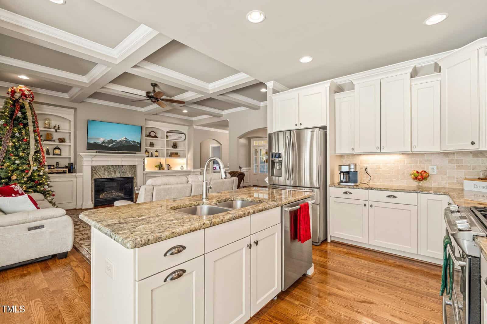 Bright kitchen with white cabinets, granite island, and stainless appliances opens to a living room with a fireplace, TV, decorated Christmas tree, and coffered ceiling. Hardwood floors throughout.