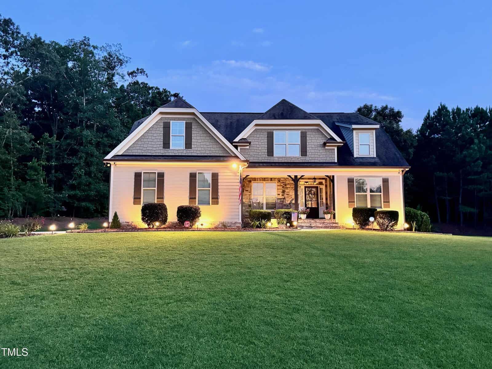 A two-story suburban house with beige siding, a dark roof, and stone accents on the front entrance, surrounded by a well-manicured lawn and illuminated by exterior ground lights at dusk. Trees are visible in the background.