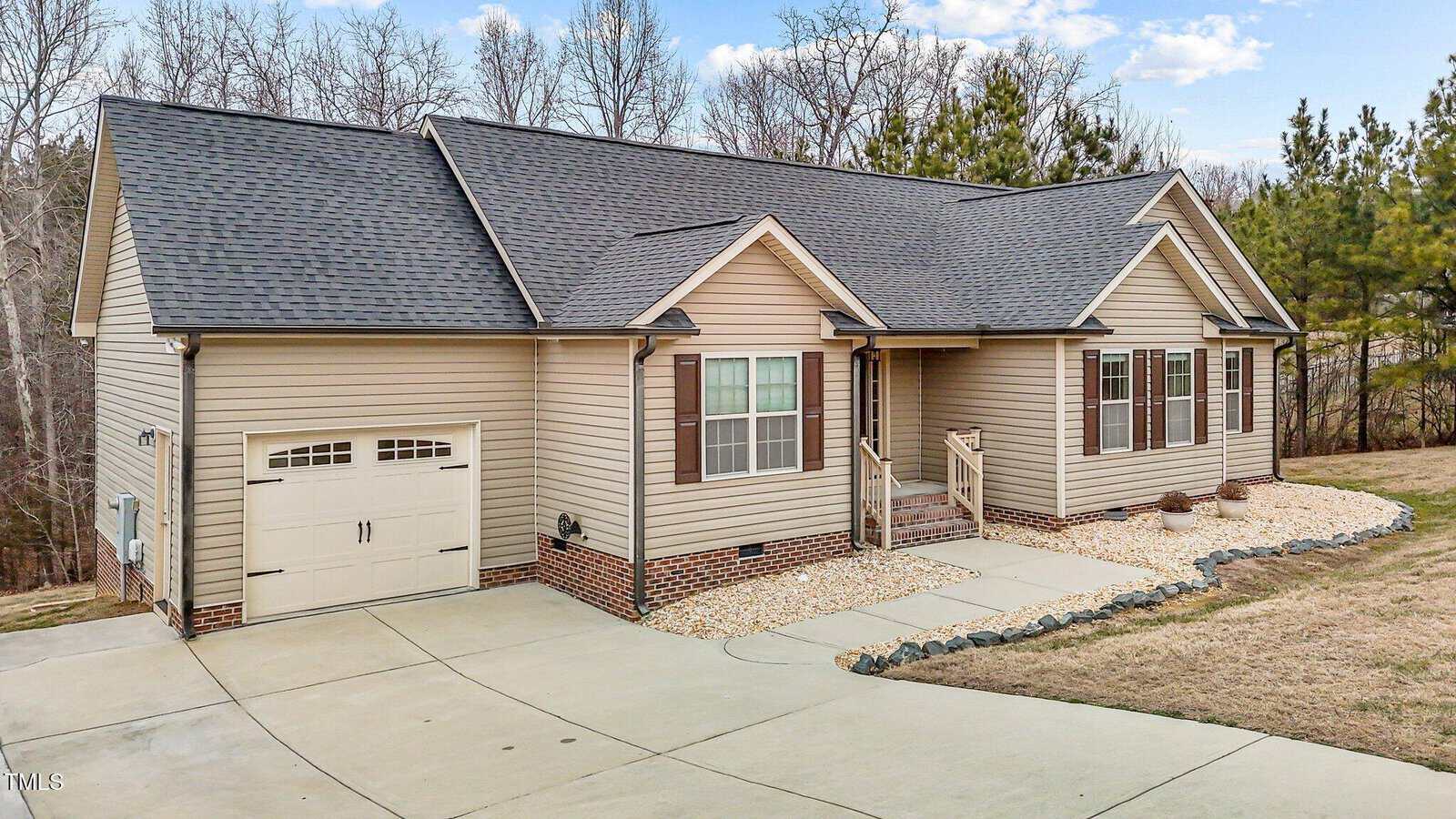 Single-story house with beige siding and a dark shingle roof. It has a front porch, a double garage door, and a curved driveway. The landscape includes a small garden with stones and shrubs, set against a backdrop of trees and a partly cloudy sky.
