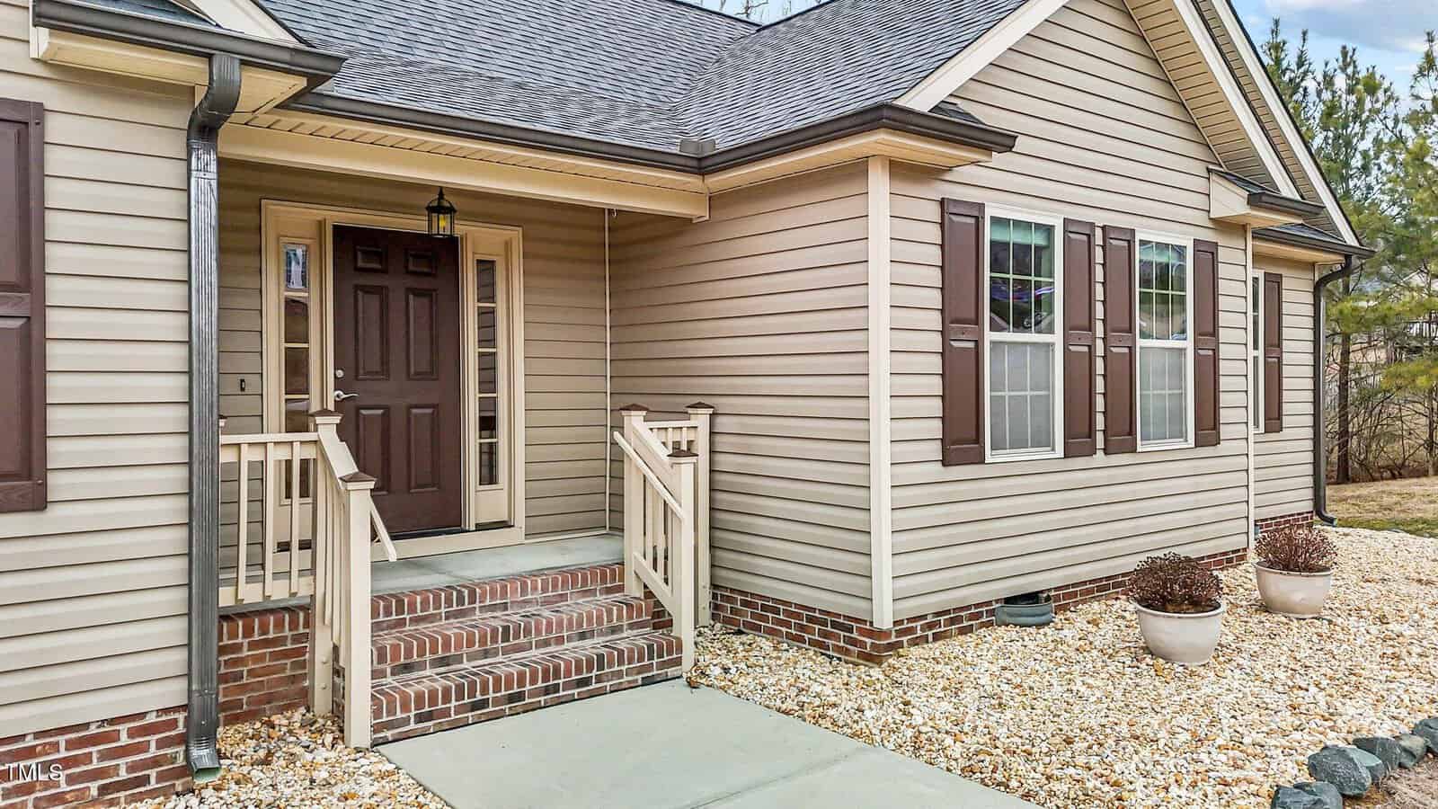 A beige house exterior with a front porch featuring a brown door and steps with railings. The porch is brick-lined at the base. There are two windows with brown shutters, and the yard is landscaped with white stones and potted plants.