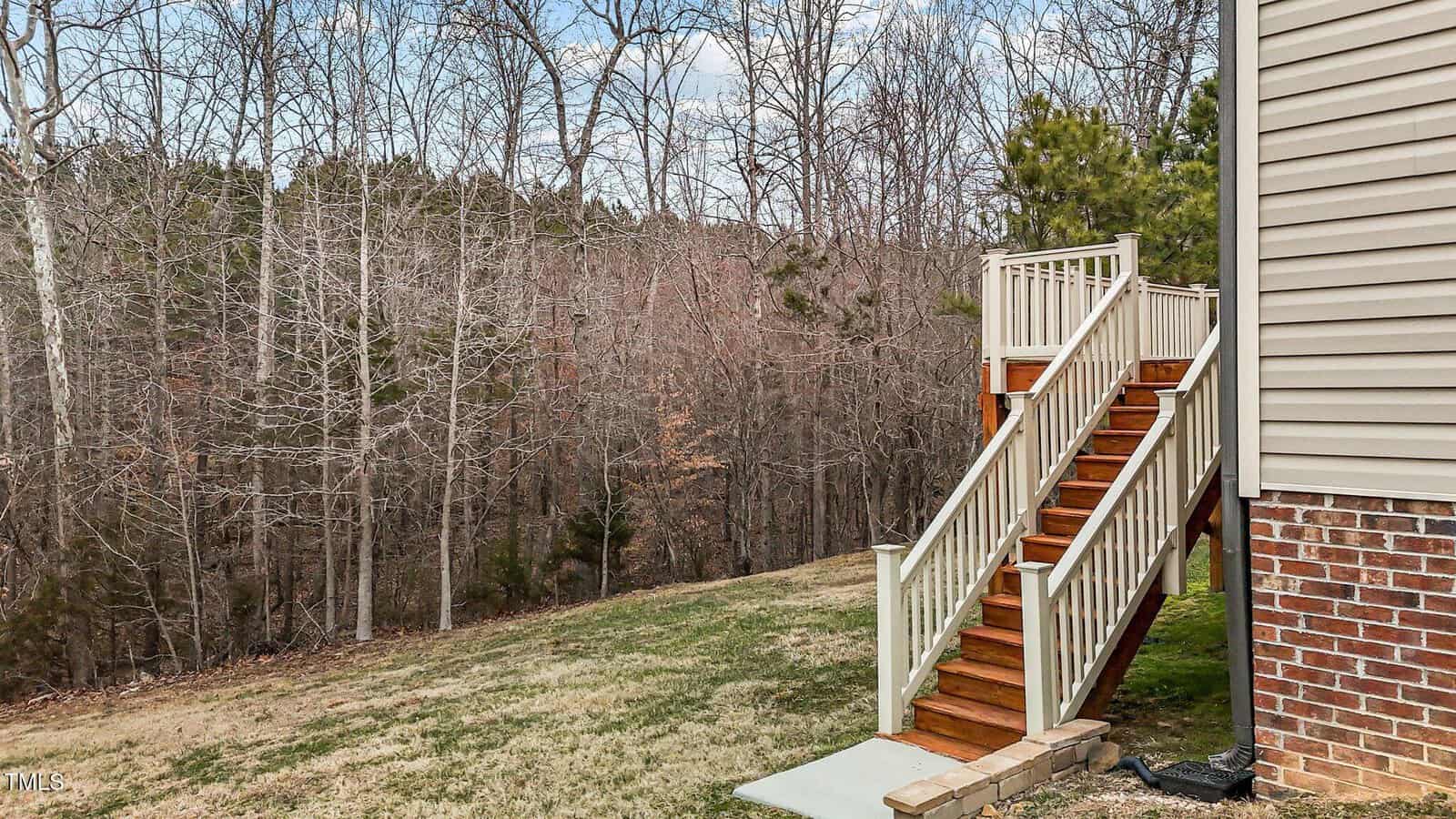 A wooden staircase with white railings leads down to a grassy backyard. The area is adjacent to the side of a brick and siding house. In the background, there is a forest with leafless trees and some evergreen trees.