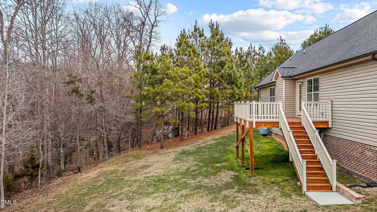 A wooden deck with stairs leads down to a grassy backyard of a house. The house is surrounded by a mix of trees, some without leaves and some evergreen. The sky is partly cloudy.