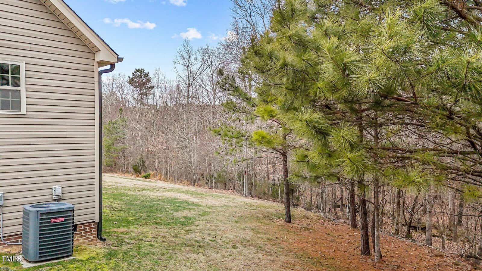 Side view of a house with beige siding and a visible air conditioning unit. The yard has green and brown grass with several pine trees on the right. In the background, there is a forested area with leafless trees. The sky is partly cloudy.