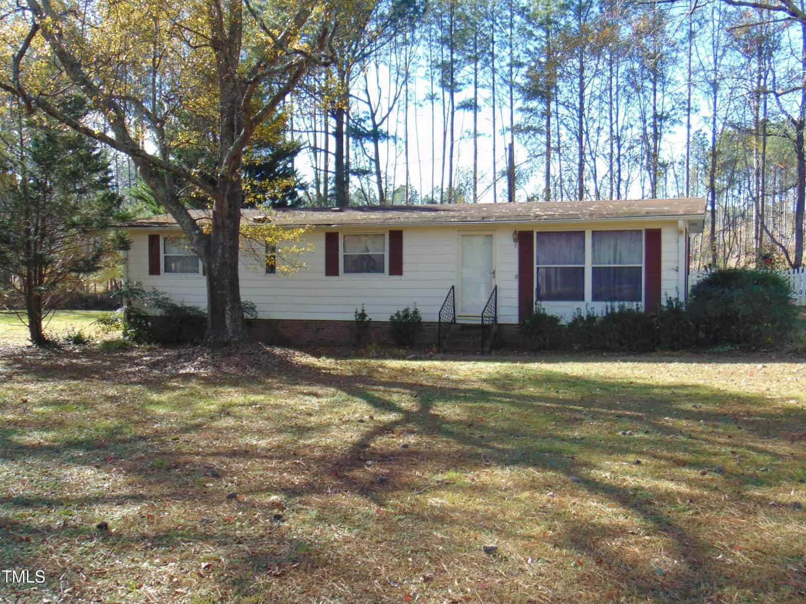 A single-story white house with brown shutters sits in front of tall trees on 1527 Cash Road, Creedmoor. It has a front porch with steps and a lawn dotted with scattered fallen leaves. A large tree casts shadows on the grass.